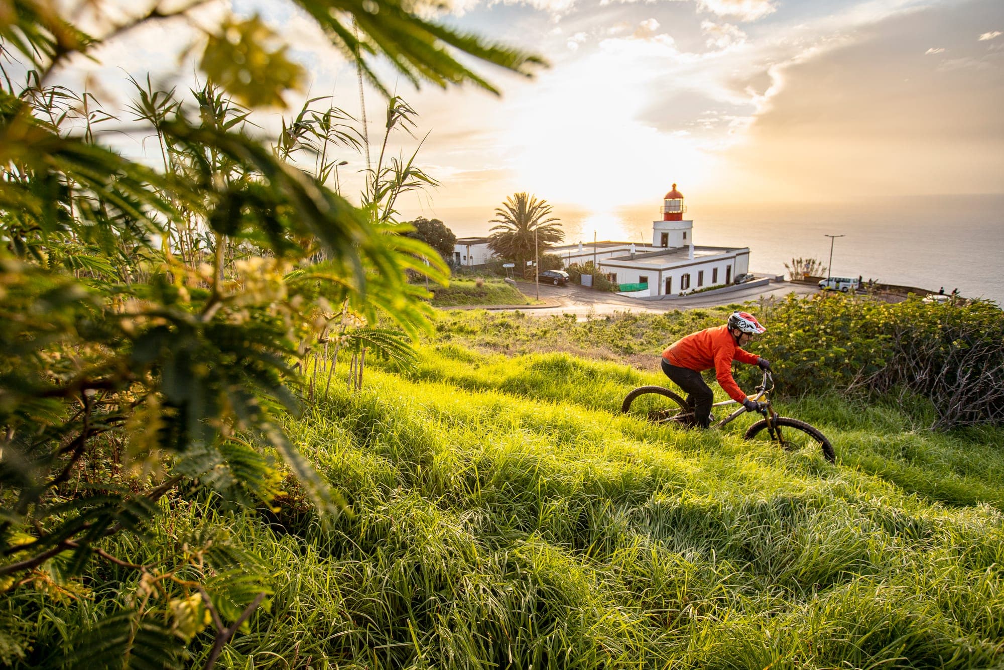 A rider on a grassy slope above a coastal lighthouse at sunset